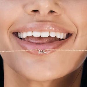 May include: A close-up of a person's mouth with a gold chain necklace with the number "444" hanging from it.