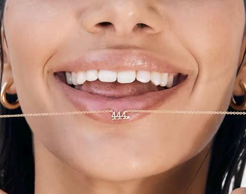 May include: A close-up of a person's mouth with a gold chain necklace with the number "444" hanging from it.