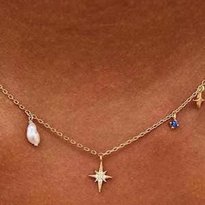 Puede incluir: Un delicado collar dorado con un colgante de estrella y varios abalorios. Los abalorios incluyen una perla blanca, una pequeña gema azul y dos estrellas doradas. El collar se lleva alrededor del cuello.