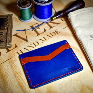 May include: A handmade blue leather cardholder with an orange accent and red stitching. The cardholder is displayed with leatherworking tools, spools of thread, and a canvas bag. The bag is printed with "SEVERS HANDMADE" and "MADE IN THE U.S.A."