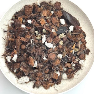 May include: A potting soil mix contained in a round, off-white bowl. The mixture consists of brown fibrous coconut husk chunks, small white perlite pieces, dark charcoal fragments, and round reddish-brown clay pebbles for plant drainage.