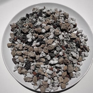 May include: A white plate filled with a mix of small, irregularly shaped rocks. The rocks vary in color, including shades of gray, brown, black, and red. The texture appears rough and porous, suggesting a natural origin.