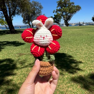 May include: A crocheted flower pot with a white bunny face and red petals. The pot is brown and has green leaves.