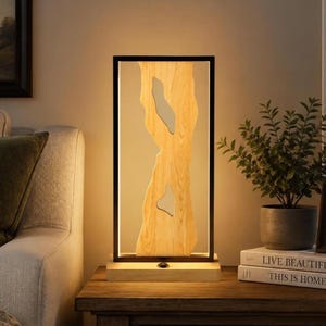 May include: A rectangular table lamp featuring a black metal frame surrounding a vertical piece of light-colored wood with two irregular cutouts. The lamp sits on a light wood base and emits a warm, soft glow from an integrated light source.