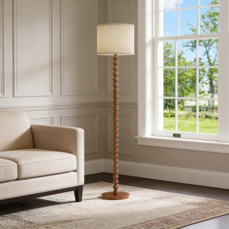 May include: A floor lamp with a tall, slender wooden stand composed of stacked spherical beads. The lamp features a cylindrical, light-colored fabric shade and a circular wooden base, standing upright on a flat surface next to a sofa.
