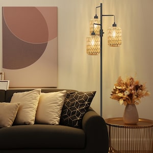 May include: A black floor lamp with three woven rattan shades, illuminating a living room scene. The lamp stands next to a dark sofa with beige pillows and a side table with a vase of dried flowers. A large abstract painting hangs on the wall.