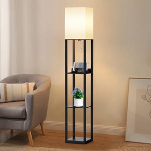 May include: A black floor lamp with a square lampshade and built-in shelves. The lamp has a rectangular frame with a light-colored lampshade. The shelves hold a tablet, a small potted plant, and other items. The lamp is next to a gray armchair.