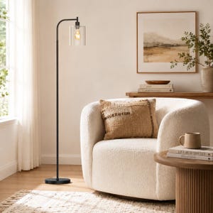 May include: A tall, slender black metal floor lamp with a single cylindrical clear glass shade and an exposed warm-toned light bulb. The lamp features a curved top arm and a circular base, providing a clean, vertical silhouette for interior lighting.