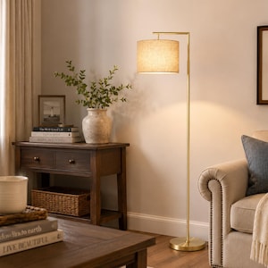 May include: A tall floor lamp with a slender, brushed gold metal stand and a circular base. The lamp features a cylindrical, textured beige fabric drum shade that emits a warm, soft glow, illuminating the surrounding space with a focused light.