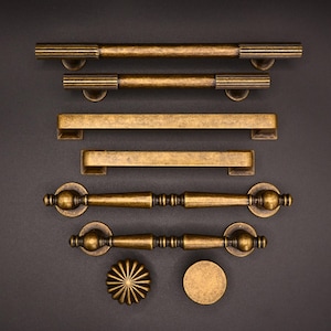 Op de afbeelding: Een set antieke messing kastbeslag, inclusief handgrepen en knoppen. De handgrepen zijn in verschillende stijlen, waaronder een lange staafgreep, een kortere staafgreep en twee handgrepen met afgeronde uiteinden. De knoppen zijn rond en hebben een decoratief ontwerp.