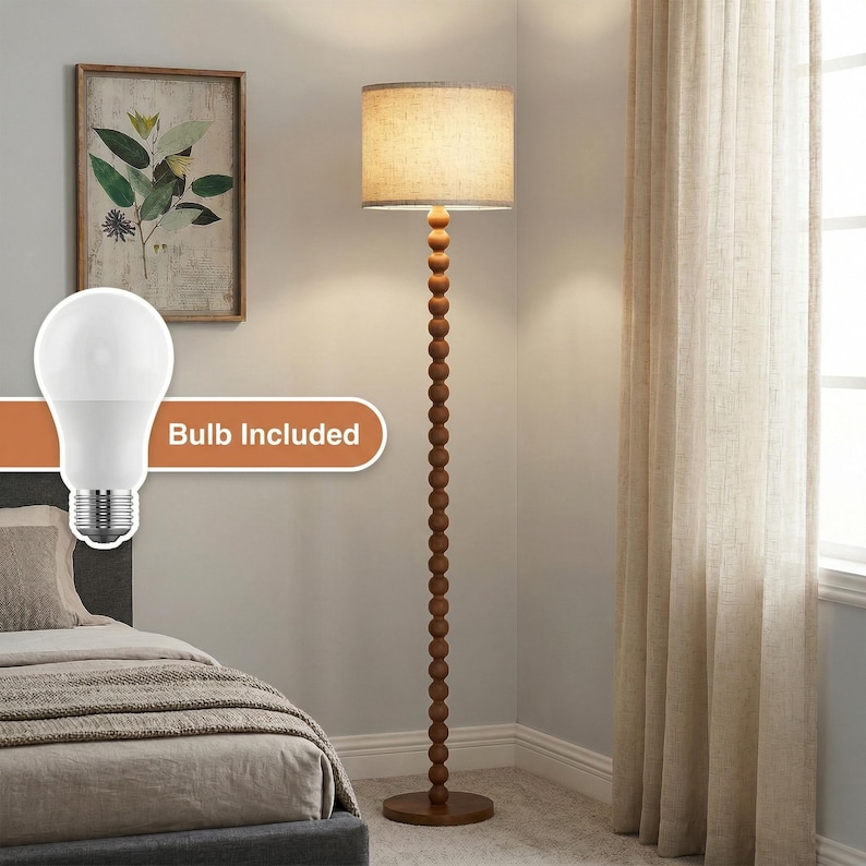 May include: A floor lamp with a tall, slender stand composed of stacked wooden spheres and a cylindrical beige fabric shade. The lamp includes a white light bulb, as indicated by the graphic overlay text reading Bulb Included.