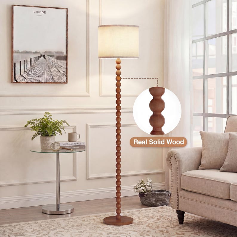 May include: A floor lamp with a tall, slender stand composed of stacked, spherical wooden beads in a medium brown finish. The lamp features a cylindrical, off-white fabric drum shade and a circular wooden base. A callout shows the wood texture.