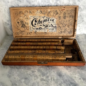 May include: A wooden box with a set of letter stamps inside. The box is labeled "Columbia Sign and Show Card Printer".