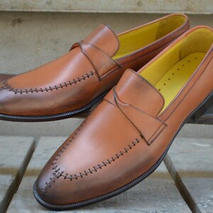 Op de afbeelding: Een paar bruine leren loafers met een gestikt detail rond de teen en een gele leren voering.