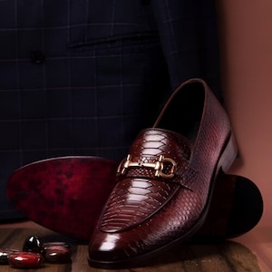 Peut inclure: Une paire de mocassins en cuir bordeaux avec un détail de boucle dorée. Les mocassins ont un motif en peau de serpent et une pointe pointue.