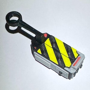 May include: A miniature Ghostbusters proton pack wand prop in black and grey, with yellow and black hazard stripes. It has a red button and a loop at the handle's end. The design is a detailed replica of the iconic movie prop.