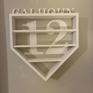 May include: White wooden baseball-shaped shelf with the number 12 and the name "CALHOUN" displayed. The shelf has multiple horizontal shelves for displaying items. The design is clean and modern, suitable for a sports-themed room.