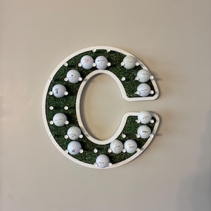 May include: A white wooden letter "C" golf ball display. The interior is covered in green turf and holds golf balls. The golf balls are white with black markings. The display is mounted on a neutral-colored wall.