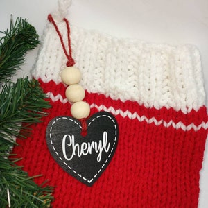 May include: Red and white knitted Christmas stocking with a black heart-shaped tag that says "Cheryl" and wooden beads hanging from the top.