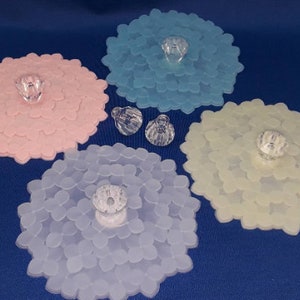 May include: Four silicone lids in different colors: pink, light blue, white, and yellow. Each lid has a flower-shaped design and a clear plastic knob in the center.