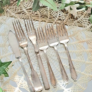 May include: A set of six silver-plated forks and a butter knife arranged on a white crocheted doily. The forks and knife have ornate handles.