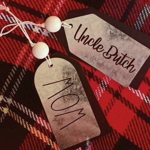 May include: Two gift tags with white wooden beads and twine. The tags are on a red and black plaid fabric. One tag says "Mom" and the other says "Uncle Butch".