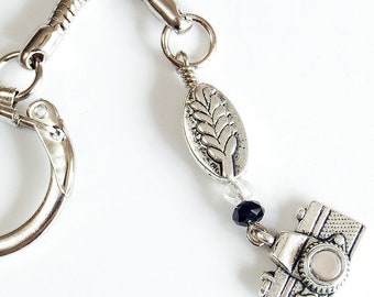 May include: Silver camera charm keychain with a black bead and a silver leaf charm. The keychain has a silver spring clasp.