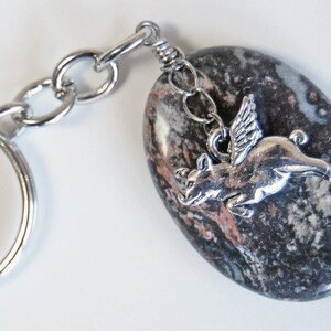 May include: A keychain featuring a polished, oval-shaped stone with a silver-colored flying pig charm. The stone is primarily gray and black with reddish-brown veining. The keychain has a silver-colored split ring and chain.