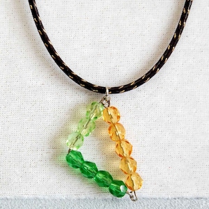 May include: A necklace with a pendant made of green and orange glass beads in the shape of a triangle. The necklace is made of black cord.