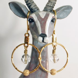 May include: Gold hoop earrings with a dangling crystal bead and a gold bead at the bottom. The earrings are displayed on a ceramic animal figurine.