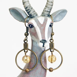 May include: A pair of gold hoop earrings with a dangling amber-colored glass bead. The earrings are hanging from the ears of a ceramic animal figurine.