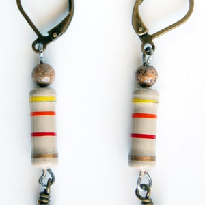 May include: A pair of earrings with brown and white beads, and beige ceramic cylinders with red and yellow stripes. The earrings have gold-toned hooks.