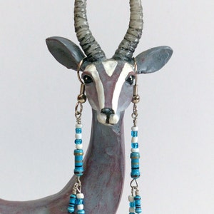 May include: A pair of long, dangling earrings with blue and white beads. The earrings are hanging from the ears of a ceramic animal figurine that resembles an antelope.