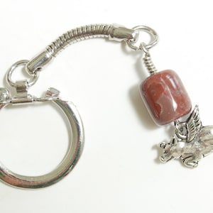 May include: A silver-toned keychain featuring a flying pig charm with wings, a cylindrical red-brown bead, and a key ring. The keychain has a decorative, flexible metal connector.