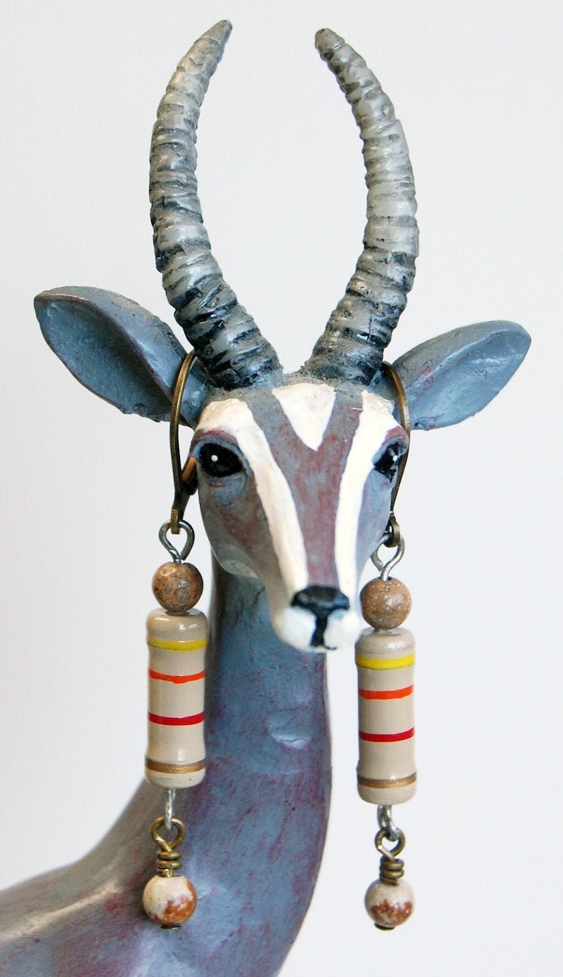 May include: A ceramic sculpture of an antelope with long, curved horns. The antelope is painted in shades of gray, white, and brown. It is wearing a pair of earrings with brown and orange beads.