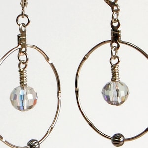 May include: A pair of silver hoop earrings with a faceted, iridescent crystal bead suspended in the center. The hoops have a hammered texture and a small, dark bead at the bottom. The earrings are designed to dangle.