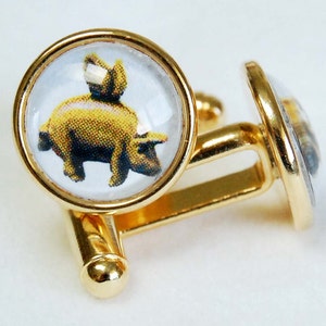 May include: Gold cufflinks with a clear glass cabochon featuring a cartoon pig. The pig is yellow and has a pink nose and hooves.