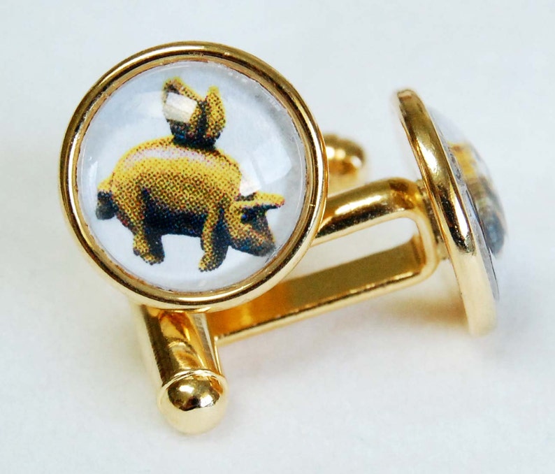 May include: Gold cufflinks with a clear glass cabochon featuring a cartoon pig. The pig is yellow and has a pink nose and hooves.