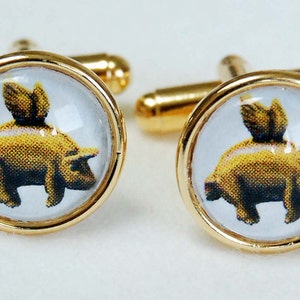 May include: A pair of gold cufflinks with a clear glass dome featuring a yellow pig with wings. The pig is facing right and has a black nose and hooves.