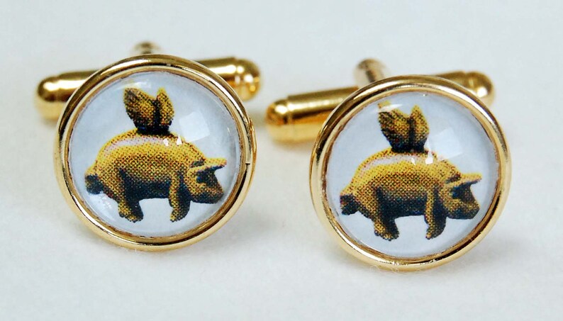 May include: A pair of gold cufflinks with a clear glass dome featuring a yellow pig with wings. The pig is facing right and has a black nose and hooves.