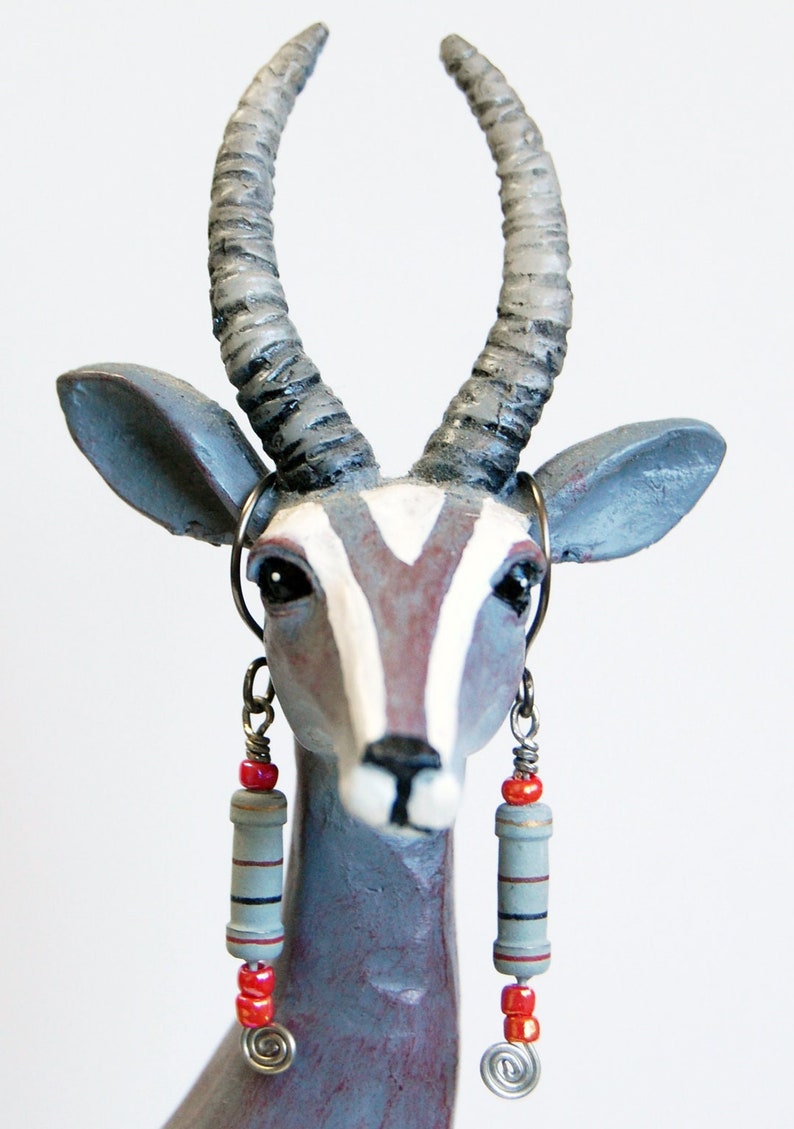 May include: A gray and white ceramic sculpture of an antelope with long, gray horns. The antelope is wearing long, dangling earrings with red and blue beads.