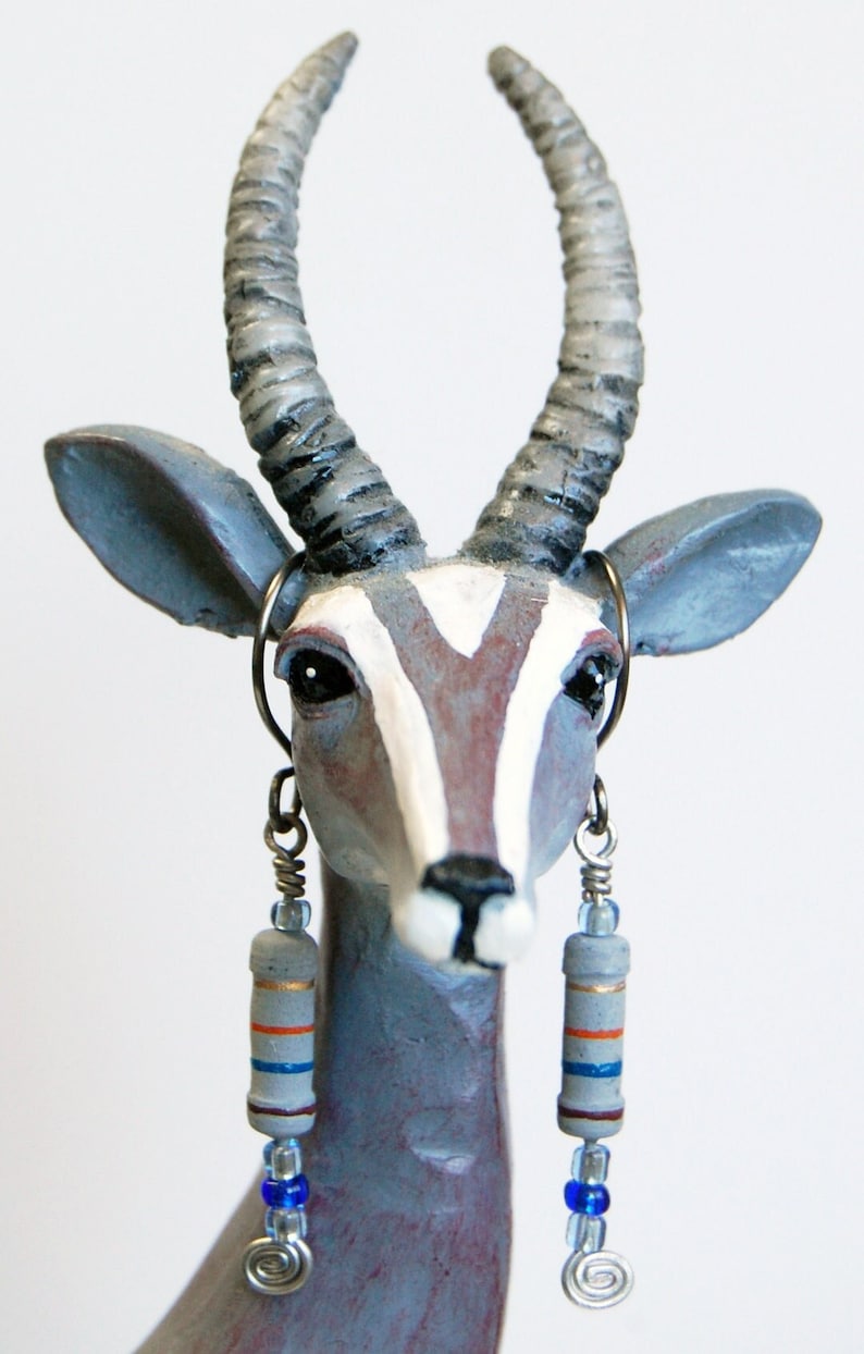 May include: A gray and white ceramic figurine of an antelope with long, twisted horns. The antelope is wearing long, dangling earrings with blue and orange beads.