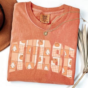 Puede incluir: Una camiseta de manga corta color óxido con la palabra NURSE en letras grandes de bloque. Las letras están rellenas con un patrón de fantasmas blancos, calabazas naranjas y cuadros. La camiseta tiene cuello redondo y una etiqueta visible.