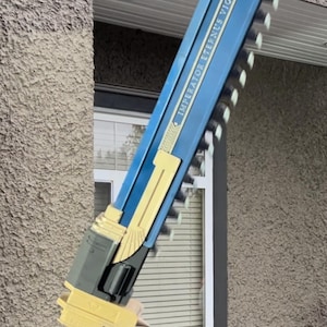 May include: A large, blue and gold prop weapon with the text "IMPERATOR AETERNUS VIGILANS". The prop has a saw-like blade and a gold handle with spikes. The weapon is held in front of a window.