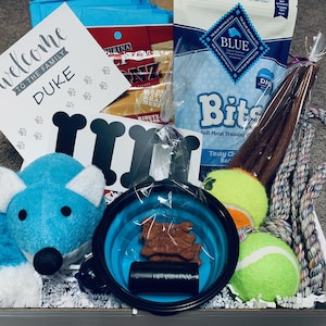 Op de afbeelding: Een welkomstcadeaupakket voor een nieuwe puppy genaamd Duke. De mand bevat een blauw pluche speelgoed, een blauwe inklapbare kom, een zakje hondenbrokjes, een zakje hondensnacks voor training, een touwspeelgoed, twee tennisballen en een bord met de tekst "Welkom in de familie Duke".