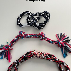 Op de afbeelding: Drie kleurrijke touwspeelgoed voor honden. Een touwspeelgoed is zwart-wit, een is rood, roze, zwart en wit, en de andere is veelkleurig. De tekst "INCLUDED" is geschreven in witte letters op een zwarte achtergrond aan de onderkant van de afbeelding. De tekst "1 Rope Toy" is geschreven in zwarte letters aan de bovenkant van de afbeelding.