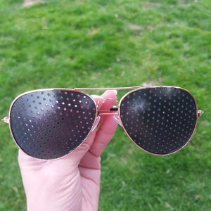 May include: Gold-framed aviator glasses with dark lenses, each dotted with numerous small holes. The glasses are held up against a background of green grass. The lenses are dark with many small holes.