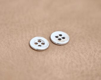 May include: Two small, round, silver buttons with four holes each, displayed on a textured, tan surface. The buttons have a shiny, metallic appearance and are evenly spaced.