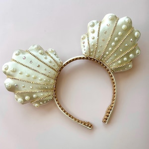 May include: A headband with seashell-shaped ears. The ears are a pale green with pearl and rhinestone accents. The headband itself is gold-colored with a braided design.