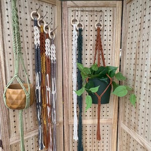Peut inclure: Plusieurs suspensions pour plantes en macramé de différentes couleurs : vert, blanc, marron et gris. L'une soutient un pot en bois, une autre un pot vert avec une plante à feuilles. Elles sont exposées sur un panneau perforé en bois.